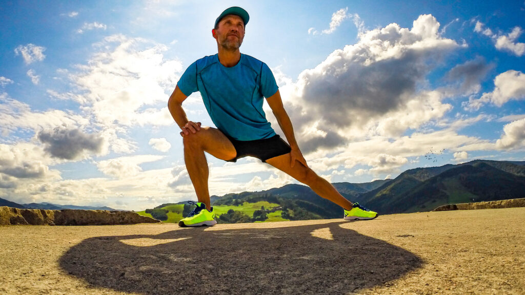 A man stands outside in the grass in front of blue sky, stretching his inner thigh by leaning to one side.