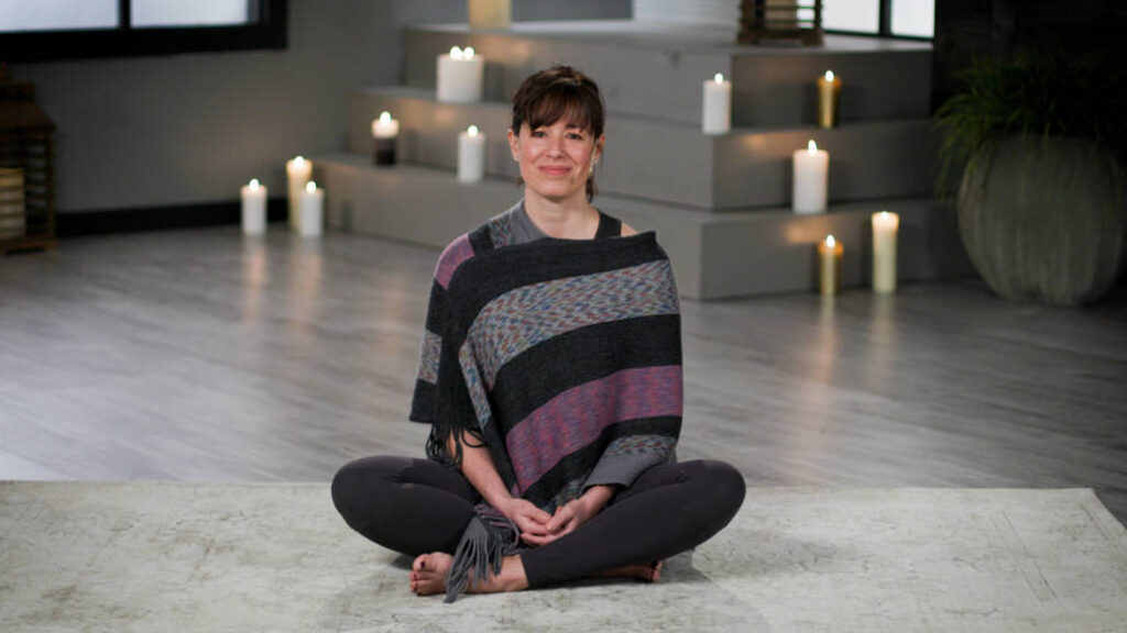 Wellbeats instructor Nicole L. leads a meditation session from a seated pose with candles lit in the background.